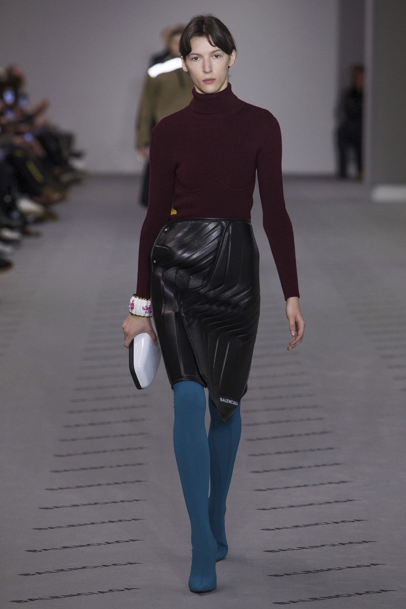 De acordo com o site da Balenciaga, essa saia de couro apareceu para o mundo no Paris Fashion Week e ela transforma o funcional em formal com estilo. Tudo isso porque ela apresenta um padrão interessante feito de couro preto e que é reforçado por neoprene, por uma estética mais utilitária. Além de todo esse visual, essa saia de couro faz parte de uma série chamada logomarca, onde o nome da marca aparece de forma assimétrica e bem proeminente. Mas, o que que foi isso tudo que eu li aqui?