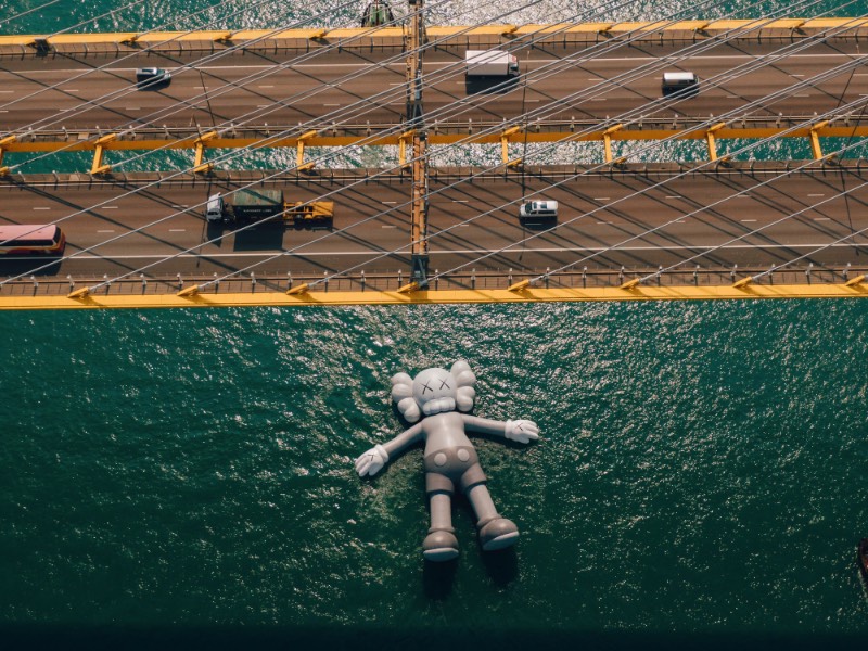 Se você estivesse olhando para o Victoria Harbour de Hong Kong, você veria uma escultura inflável criada pelo artista de rua conhecido como KAWS. Essa cena surreal é realidade, por incrível que pareça. Essa obra monocromática é a maior criação do artista americano e ela foi pensada para representar uma posição de paz, como se ela flutuasse na água olhando para as nuvens e o céu acima de nós. 