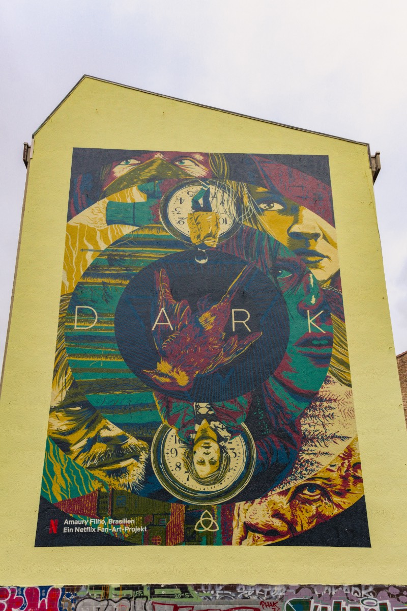 O terceiro mural de Dark em Berlim que visitei nesse domingo fica no coração de Kreuzberg, na lateral de um dos prédios próximos de Kottbusser Tor. Esse mural recebeu o nome de Sic Mundus Creatus Est e usa de vários elementos da série de forma ilustrada com uma sofisticação visual que eu não esperava ver em um mural. O mais interessante aqui é que esse é o trabalho de um brasileiro chamado Amaury Filho. Carioca, designer e ilustrador que mora em Curitiba há quase 15 anos e trabalha com o mercado publicitário na capital paranaense. Além de ter feito o meu mural de Dark favorito.