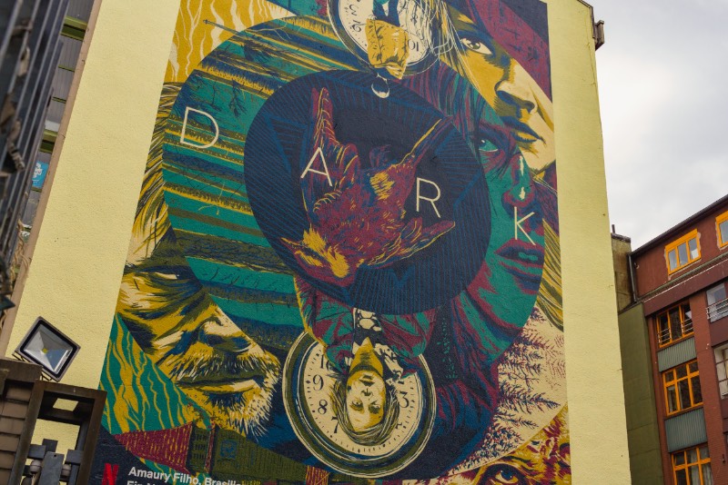 O terceiro mural de Dark em Berlim que visitei nesse domingo fica no coração de Kreuzberg, na lateral de um dos prédios próximos de Kottbusser Tor. Esse mural recebeu o nome de Sic Mundus Creatus Est e usa de vários elementos da série de forma ilustrada com uma sofisticação visual que eu não esperava ver em um mural. O mais interessante aqui é que esse é o trabalho de um brasileiro chamado Amaury Filho. Carioca, designer e ilustrador que mora em Curitiba há quase 15 anos e trabalha com o mercado publicitário na capital paranaense. Além de ter feito o meu mural de Dark favorito.