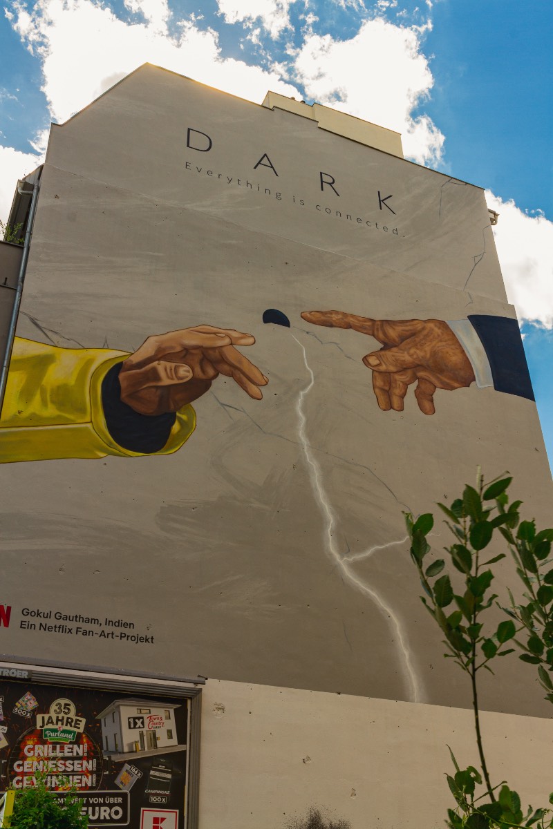 O primeiro mural de Dark em Berlim que eu visitei foi em Prenzlauer Berg, em uma das avenidas que liga o norte da cidade com o centro. Foi lá que eu me deparei com o trabalho de Gokul Gautham que, parece, ter usado de uma referência uma das pinturas mais clássicas da Capela Cistina. Mas, em seu mural, temos as mãos de Jonas e Adam se aproximando perto do que me parece ser uma versão simplificada da caverna. 