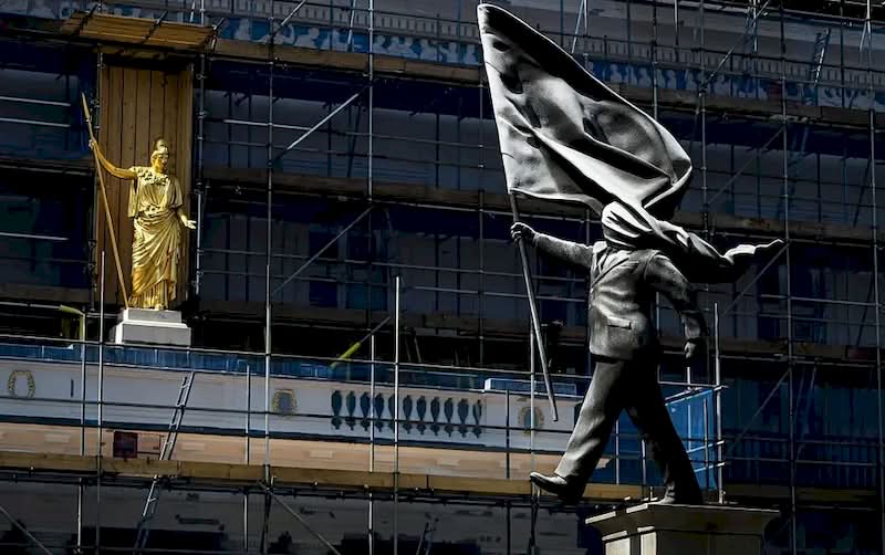 A histórica Waterloo Place, em Londres, é conhecida por seus monumentos a heróis de guerra e à realeza. Mas, no final de abril de 2026, as pessoas que passaram por essa praça foram surpreendidas com uma nova adição à paisagem urbana. Uma estátua do Banksy foi colocada em um pedestal, retratando um homem de terno, cego pela bandeira que carrega, congelado no instante em que parece estar à beira de um precipício.