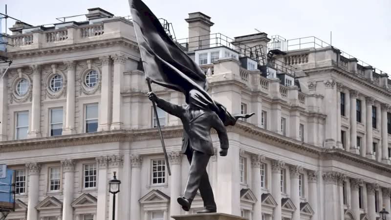 A histórica Waterloo Place, em Londres, é conhecida por seus monumentos a heróis de guerra e à realeza. Mas, no final de abril de 2026, as pessoas que passaram por essa praça foram surpreendidas com uma nova adição à paisagem urbana. Uma estátua do Banksy foi colocada em um pedestal, retratando um homem de terno, cego pela bandeira que carrega, congelado no instante em que parece estar à beira de um precipício.