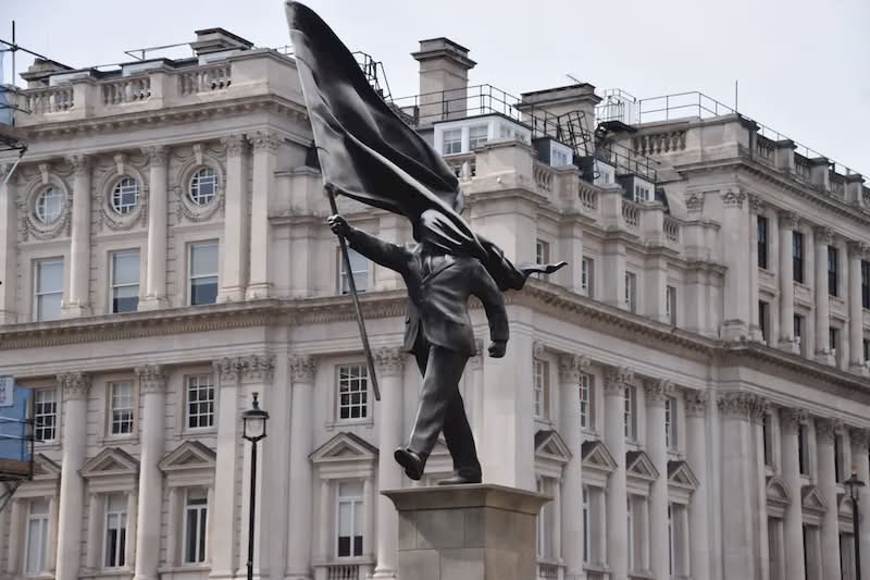 A histórica Waterloo Place, em Londres, é conhecida por seus monumentos a heróis de guerra e à realeza. Mas, no final de abril de 2026, as pessoas que passaram por essa praça foram surpreendidas com uma nova adição à paisagem urbana. Uma estátua do Banksy foi colocada em um pedestal, retratando um homem de terno, cego pela bandeira que carrega, congelado no instante em que parece estar à beira de um precipício.